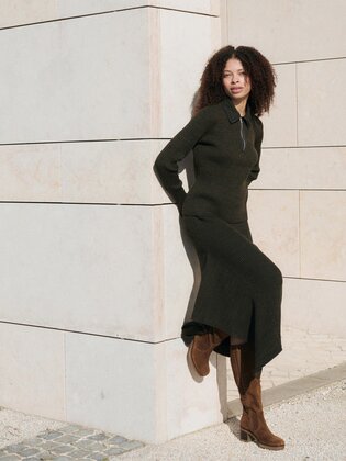 En kvinna står avslappnat vid en ljus stenmur, har på sig en mörk lång klänning och bruna stövlar. Bilden är tagen utomhus i dagsljus. | © Gabor Shoes GmbH, Tyskland