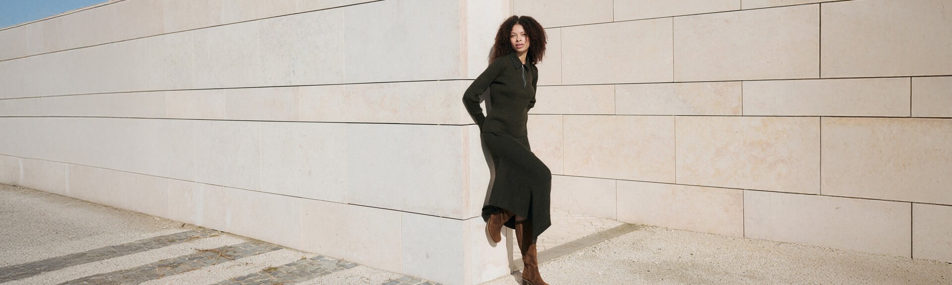 En kvinna står avslappnat vid en ljus stenmur, har på sig en mörk lång klänning och bruna stövlar. Bilden är tagen utomhus i dagsljus. | © Gabor Shoes GmbH, Tyskland