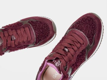En kvinna med beige byxor bär vinröda sneakers och står framför två speglar så att skorna reflekteras flera gånger. | © Gabor Shoes GmbH, Tyskland