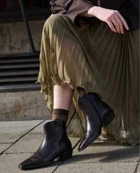 Eine Frau trägt dunkelbraune Damen-Stiefeletten mit spitzer Kappe und kleinem Absatz zu einem langen, fließenden olivgrünen Rock; sie steht auf einem gepflasterten Gehweg vor Steinstufen. | © Gabor Shoes GmbH, Rosenheim