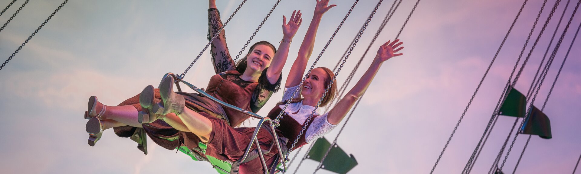 Zwei Frauen sitzen nebeneinander auf einem Kettenkarussell und strecken fröhlich die Arme in die Luft. Sie tragen traditionelle Dirndl und das Bild zeigt den Himmel im Hintergrund. | © Gabor Shoes GmbH, Rosenheim