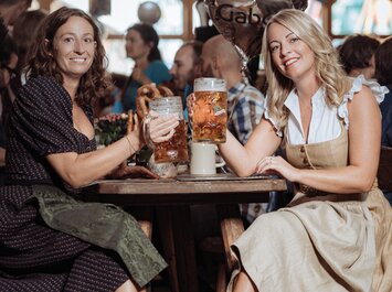 Zwei Frauen in Dirndl stoßen in einem Festzelt mit großen Biergläsern an. Auf dem Tisch stehen Brezeln und Blumen, im Hintergrund sind weitere Gäste und ein Herzluftballon mit „Gabor“-Schriftzug zu sehen. | © Gabor Shoes GmbH, Rosenheim