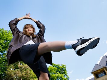 Eine Frau steht im Freien, trägt eine dunkle Jacke, schwarzen Rock und schwarze Sneaker, ein Bein ist in die Luft gestreckt. | © Gabor Shoes GmbH, Rosenheim
