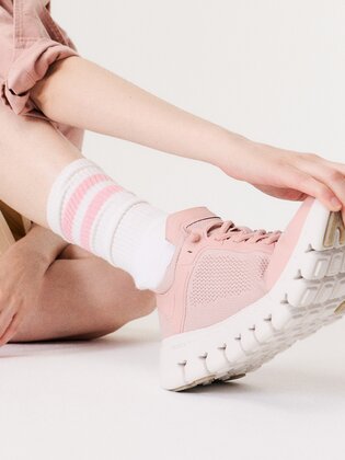 En kvinna sitter på golvet och bär rosa sneakers med vita och rosa strumpor. Hennes hand rör vid skosulan, bakgrunden är vit och enkel. | © Gabor Shoes GmbH, Tyskland