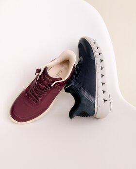 Eine Frau sitzt auf dem Boden in einem hellen Raum und trägt rosa Sneaker, gestreifte Socken, einen cremefarbenen Rock und eine rosa Jacke. | © Gabor Shoes GmbH, Rosenheim
