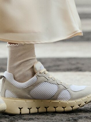Due gambe con calzini sportivi bianchi e sneakers color crema sono su un passaggio pedonale. La donna indossa una gonna lunga e chiara. | © Gabor Shoes GmbH, Germania