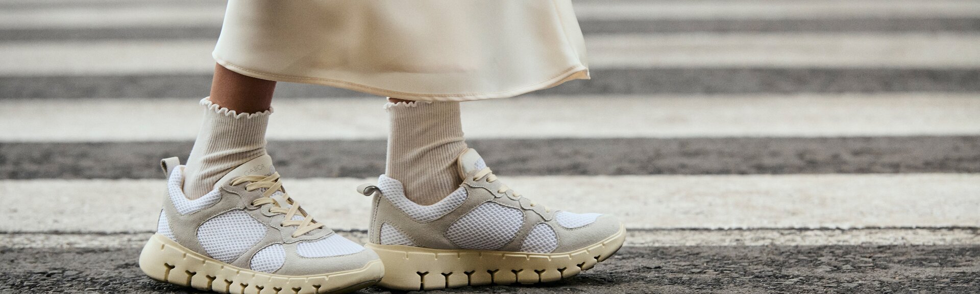 Två ben i vita sportstrumpor och krämfärgade sneakers står på ett övergångsställe. Kvinnan bär en lång, ljus kjol. | © Gabor Shoes GmbH, Tyskland
