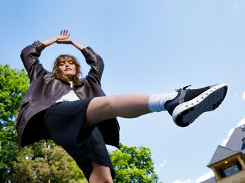 Eine Frau im schwarzen Outfit und mit sportlichen Schuhen streckt ein Bein nach vorne und steht vor grünem Parkhintergrund. | © Gabor Shoes GmbH, Rosenheim