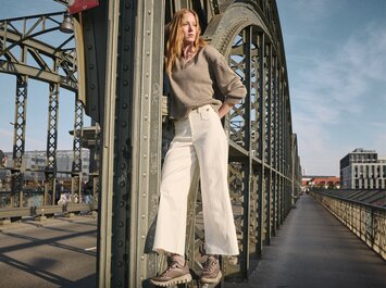 Una donna è in piedi in modo informale davanti a un muro di pietra chiaro, indossa pantaloni grigi larghi, sneakers azzurri, una camicia lunga a righe blu e ha un maglione giallo legato in modo morbido sui fianchi. Fotografia dal basso. | © Gabor Shoes GmbH, Germania