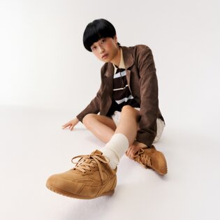 En kvinna sitter på golvet i ett vitt rum och bär bruna sneakers, vita strumpor, en beige kjol, en randig topp och en brun kavaj. | © Gabor Shoes GmbH, Tyskland