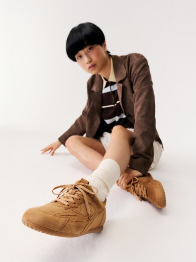 En kvinna sitter på golvet i ett vitt rum och bär bruna sneakers, vita strumpor, en beige kjol, en randig topp och en brun kavaj. | © Gabor Shoes GmbH, Tyskland