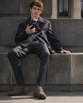 Junger Mann sitzt entspannt auf einer Steinbank, hört mit Kopfhörern Musik auf seinem Smartphone und trägt einen dunkelgrauen Anzug mit braunen Gabor-Schnürboots. | © Gabor Shoes GmbH, Rosenheim