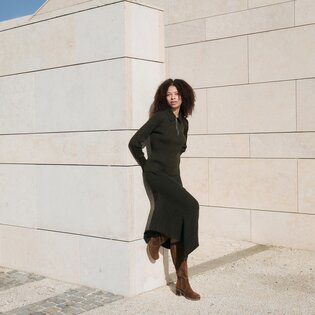 Eine Frau mit lockigen Haaren lehnt an einer hellen Steinwand, trägt ein langes schwarzes Kleid und braune Stiefel. Die Szene ist sonnig und ruhig. | © Gabor Shoes GmbH, Rosenheim