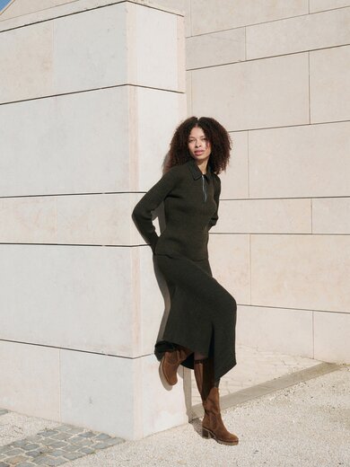 En kvinna med lockigt hår lutar mot en ljus stenmur, har på sig en lång svart klänning och bruna stövlar. Scenen är solig och lugn. | © Gabor Shoes GmbH, Tyskland