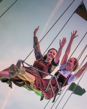 Zwei Frauen sitzen nebeneinander auf einem Kettenkarussell und strecken fröhlich die Arme in die Luft. Sie tragen traditionelle Dirndl und das Bild zeigt den Himmel im Hintergrund. | © Gabor Shoes GmbH, Rosenheim