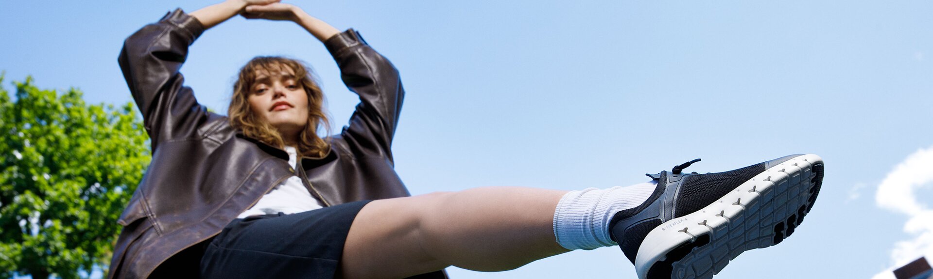 En kvinna står utomhus, har på sig en mörk jacka, svart kjol och svarta sneakers, ett ben är lyft upp i luften. | © Gabor Shoes GmbH, Tyskland