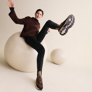 Una donna assume una posa dinamica in uno skatepark, indossa una camicia azzurra, una gonna bianca e sneakers beige. | © Gabor Shoes GmbH, Germania