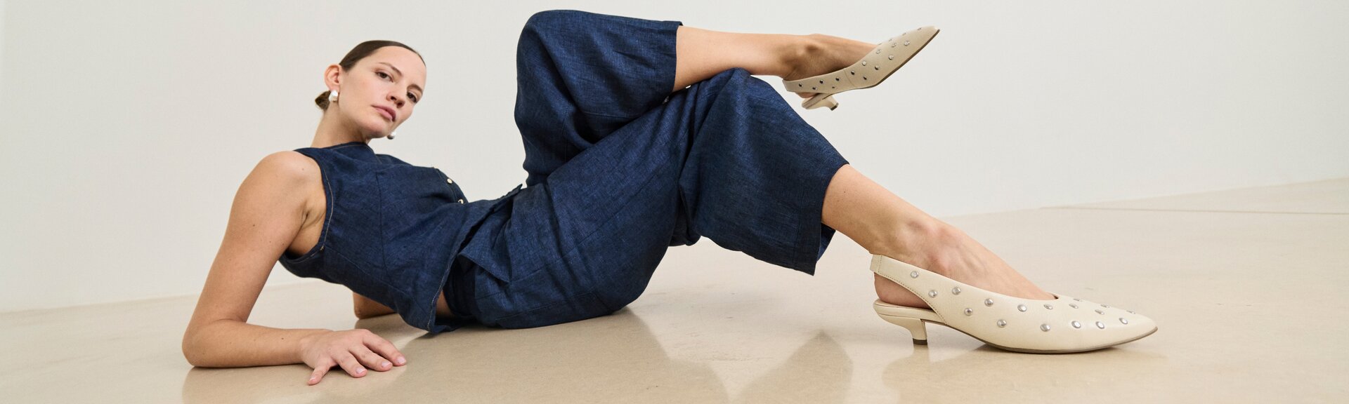 En kvinna har på sig en mörkblå jumpsuit och ligger avslappnat på golvet. De beige pumpsen med små silvernitar och låg klack är bildens fokus. | © Gabor Shoes GmbH, Tyskland