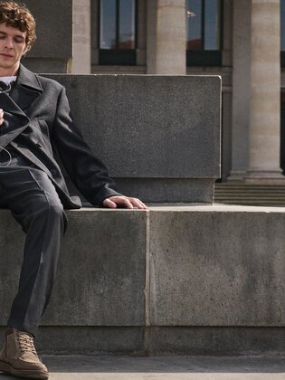 Jeune homme assis détendu sur un banc en pierre, écoutant de la musique avec des écouteurs sur son smartphone et portant un costume gris foncé avec des bottines à lacets Gabor marron. | © Gabor Shoes GmbH, Allemagne