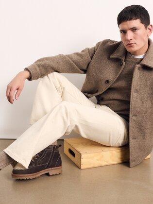 Ein Mann sitzt auf einem Holzklotz vor einer weißen Wand und trägt helle Hose, braunen Pullover, hellbraune Jacke und dunkle Schnürboots. | © Gabor Shoes GmbH, Rosenheim
