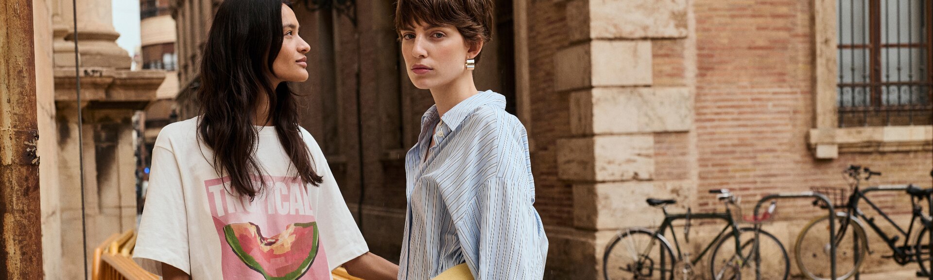 Två kvinnor står bredvid varandra framför en gammal byggnad; den ena har på sig en vit t-shirt med vattenmelontryck och texten "Tropical", den andra en ljusblå skjorta. I bakgrunden syns cyklar lutade mot ett räcke. | © Gabor Shoes GmbH, Tyskland