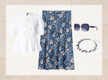 En outfit består av en vit blus med korta puffärmar, en knälång kjol med blåvitt bladmotiv, mörkblå solglasögon och ett matchande blått halsband. Allt ligger bredvid varandra mot en ljus bakgrund. | © aboutyou.de 
