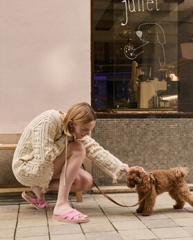 Eine Frau in Strickkleid und rosa Pantoletten kniet auf einem Gehweg und streichelt einen kleinen braunen Hund an der Leine. Im Hintergrund eine Bank und ein Schaufenster mit der Aufschrift "juliet" sowie einer gezeichneten Hundegrafik. | © Gabor Shoes GmbH, Rosenheim