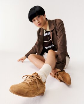 Eine Frau sitzt auf dem Boden in einem weißen Raum und trägt braune Sneaker, weiße Socken, einen beigen Rock, ein gestreiftes Oberteil und einen braunen Blazer. | © Gabor Shoes GmbH, Rosenheim