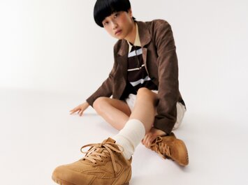 En kvinna sitter på golvet i ett vitt rum och bär bruna sneakers, vita strumpor, en beige kjol, en randig topp och en brun kavaj. | © Gabor Shoes GmbH, Tyskland