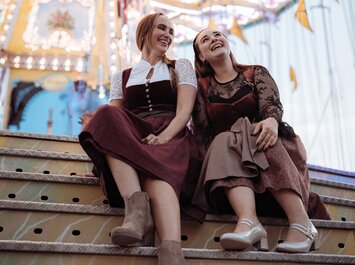 Zwei Frauen sitzen nebeneinander auf einer Treppe vor einem bunten Karussell. Sie tragen festliche Dirndlkleider und Absatzschuhe. Die Stimmung wirkt entspannt und fröhlich. | © Gabor Shoes GmbH, Rosenheim