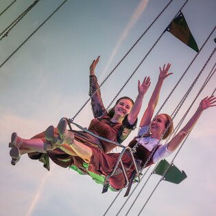Zwei Frauen sitzen nebeneinander auf einem Kettenkarussell und strecken fröhlich die Arme in die Luft. Sie tragen traditionelle Dirndl und das Bild zeigt den Himmel im Hintergrund. | © Gabor Shoes GmbH, Rosenheim