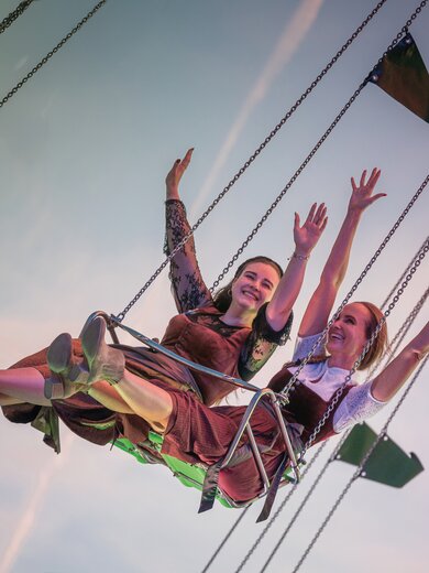Zwei Frauen sitzen nebeneinander auf einem Kettenkarussell und strecken fröhlich die Arme in die Luft. Sie tragen traditionelle Dirndl und das Bild zeigt den Himmel im Hintergrund. | © Gabor Shoes GmbH, Rosenheim