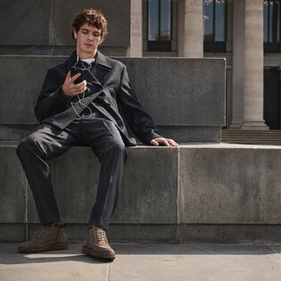 Junger Mann sitzt entspannt auf einer Steinbank, hört mit Kopfhörern Musik auf seinem Smartphone und trägt einen dunkelgrauen Anzug mit braunen Gabor-Schnürboots. | © Gabor Shoes GmbH, Rosenheim