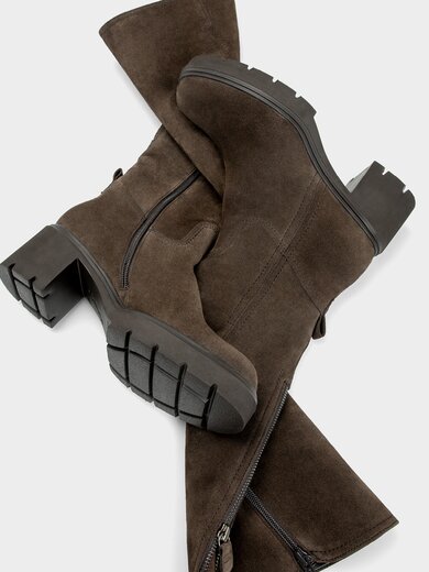 Een vrouw zit ontspannen in elegante bruine kleding en bruine laarzen op een zanderige ondergrond tussen duinen en gras. Op de achtergrond zijn groene struiken en bomen te zien. | © Gabor Shoes GmbH, Duitsland