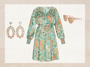 En färgglad klänning med långa ärmar i grönt och orange står i fokus. Till vänster finns iögonfallande ovala örhängen med glittrande stenar, till höger ett par beige solglasögon med bred ram. Alla föremål syns mot ljus bakgrund. | © aboutyou.de 