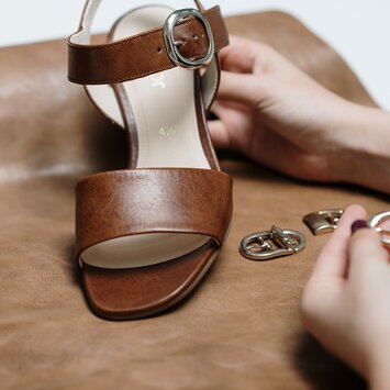 Braune Sandalette mit Metallschnalle liegt auf Leder, Hände halten Schnallen. | © Gabor Shoes GmbH, Rosenheim