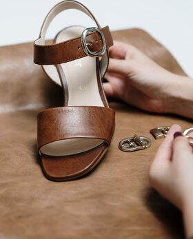 Braune Sandalette mit Metallschnalle liegt auf Leder, Hände halten Schnallen. | © Gabor Shoes GmbH, Rosenheim
