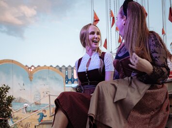 Två kvinnor sitter bredvid varandra på en trappa framför en färgglad karusell. De har festliga dirndlklänningar och klackskor. Stämningen är avslappnad och glad. | © Gabor Shoes GmbH, Tyskland