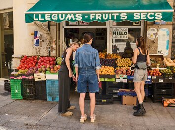  Devant un petit stand de fruits et légumes avec un auvent vert et l’inscription « LLEGUMS • FRUITS SECS » se tiennent plusieurs personnes. Sur des tables et dans des caisses sont disposés différents fruits et légumes colorés. Le magasin se trouve à un coin de rue. | © Gabor Shoes GmbH, Allemagne