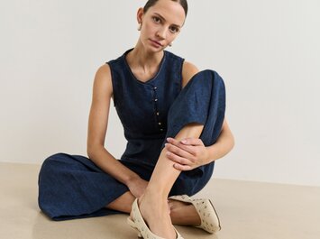 Eine Frau trägt einen dunkelblauen Jumpsuit und sitzt entspannt auf dem Boden. Sie trägt beigefarbene Pumps mit kleinen silbernen Nieten und kleinem Absatz. | © Gabor Shoes GmbH, Rosenheim