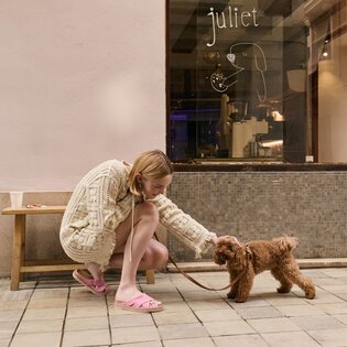 En kvinna i beige stickad klänning och rosa slip-in skor knäböjer på en trottoar och klappar en liten brun hund i koppel. I bakgrunden syns en bänk och ett skyltfönster med texten "juliet" samt en tecknad hundgrafik. | © Gabor Shoes GmbH, Tyskland