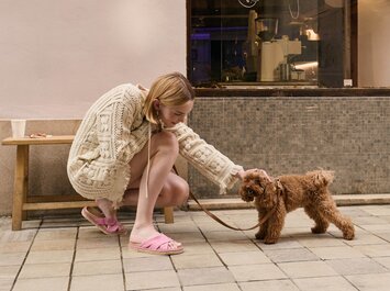 En kvinna i beige stickad klänning och rosa slip-in skor knäböjer på en trottoar och klappar en liten brun hund i koppel. | © Gabor Shoes GmbH, Tyskland