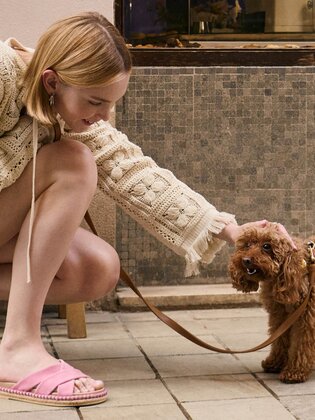 Eine Frau in einem beigen Strickkleid und rosa Pantoletten kniet auf dem Gehweg und streichelt einen kleinen braunen Hund mit lockigem Fell. Daneben stehen eine Holzbank mit einem Becher darauf und ein mintfarbenes Fahrrad. | © Gabor Shoes GmbH, Rosenheim