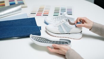 Zwei weiße Sneaker mit Stoffmustern und Farbpaletten auf einem Tisch, Hände halten Schuh und Sohle. | © Gabor Shoes GmbH, Rosenheim