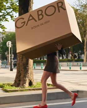 Gabor Damenschuhe | Eine Frau läuft durch die Stadt in roten Gabor Ballerinas und trägt einen übergroßen, braunen Schuhkarton mit dem Gabor Logo und der Aufschrift "Born in 1919". | © Gabor Shoes GmbH, Rosenheim