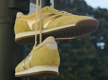 Gabor Damenschuhe | Gelbe Gabor-Sneaker mit weißen Sohlen und metallisch glänzenden Streifen, hängen an ihren Schnürsenkeln vor einem unscharfem Hintergrund. | © Gabor Shoes GmbH, Rosenheim