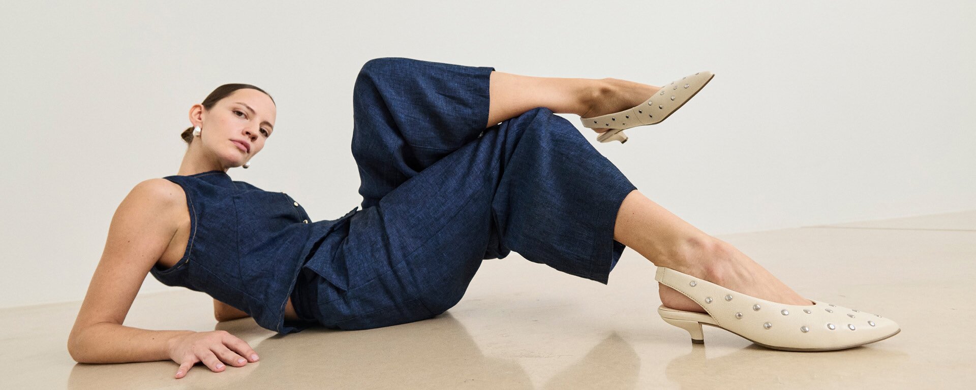 Gabor Damenschuhe | Eine Frau liegt auf hellem Boden, trägt einen dunkelblauen Jumpsuit und cremefarbene Slingpumps mit Nieten von Gabor. | © Gabor Shoes GmbH, Rosenheim