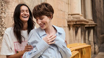 myGabor | Twee lachende vrouwen staan iets verspringend achter elkaar voor een oude stenen muur, close-up van bovenlichaam en gezichten | © Gabor Shoes GmbH, Rosenheim