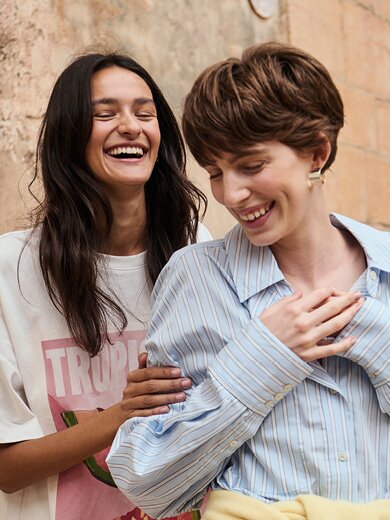 myGabor | Twee lachende vrouwen staan iets verspringend achter elkaar voor een oude stenen muur, close-up van bovenlichaam en gezichten | © Gabor Shoes GmbH, Rosenheim