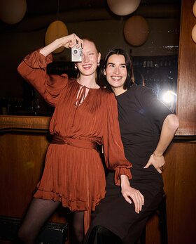 Zwei Frauen stehen eng beieinander in einer Bar, lächeln in die Kamera, eine hält eine kleine Fotokamera hoch. | © Gabor Shoes GmbH, Rosenheim